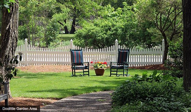 A peaceful backyard garden with two chairs facing each other next to a white picket fence.