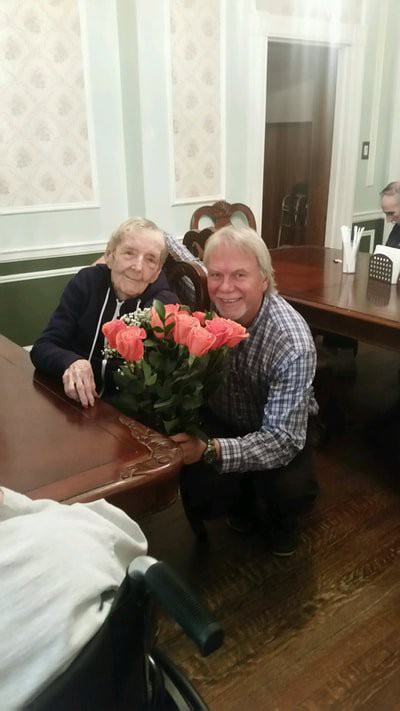 A man presents a bouquet of flowers to an elderly woman at a table indoors.