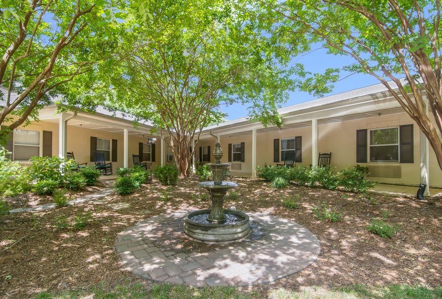 A serene courtyard featuring a central fountain surrounded by lush trees and a building with large windows.