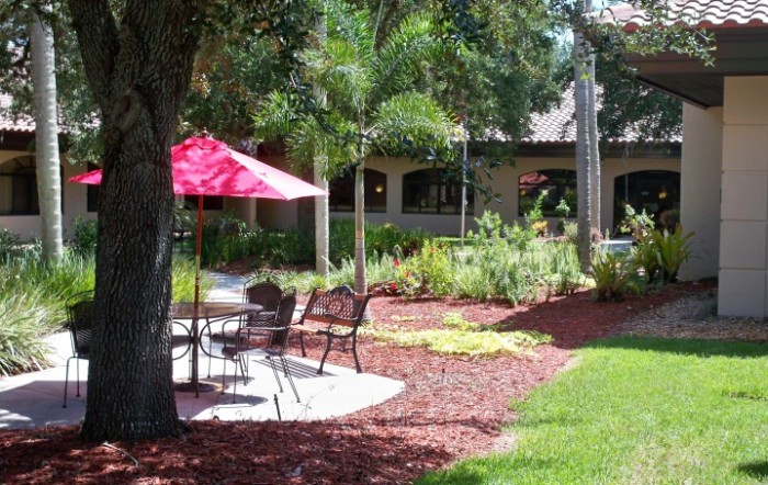 A peaceful garden patio with chairs and a red umbrella, surrounded by lush greenery.