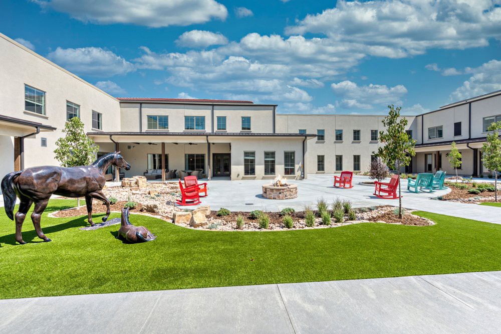 A modern building with a landscaped courtyard featuring red chairs, sculptures, and greenery under a sunny sky.