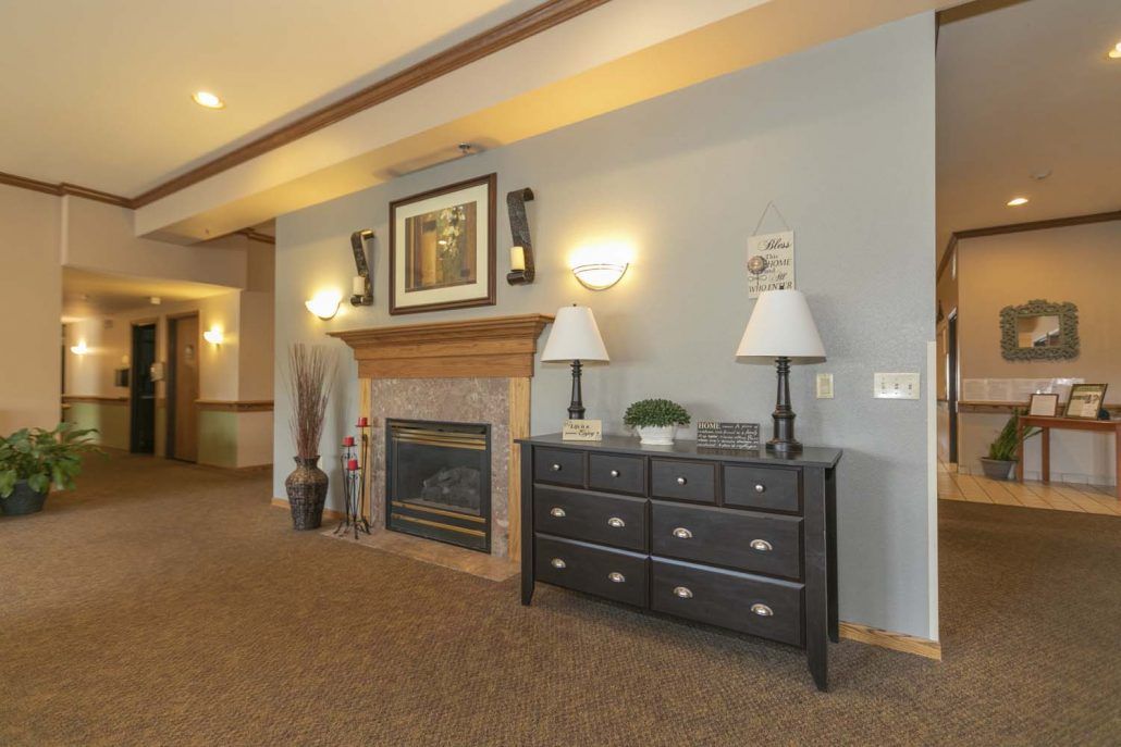 A cozy living room interior featuring a fireplace, a chest of drawers with two lamps, and wall artwork.