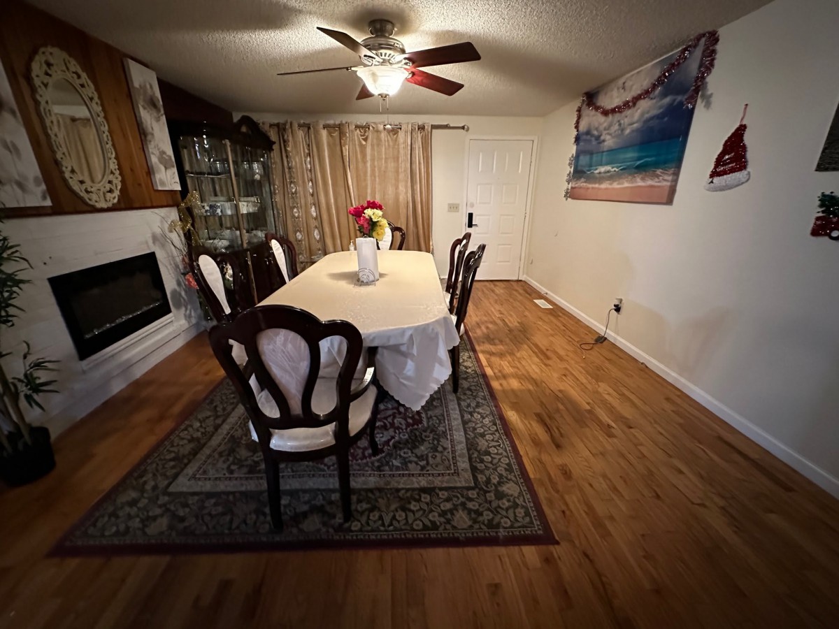 A cozy dining room with a table set for six, featuring a ceiling fan, artwork on the wall, and a decorative rug on wooden flooring.
