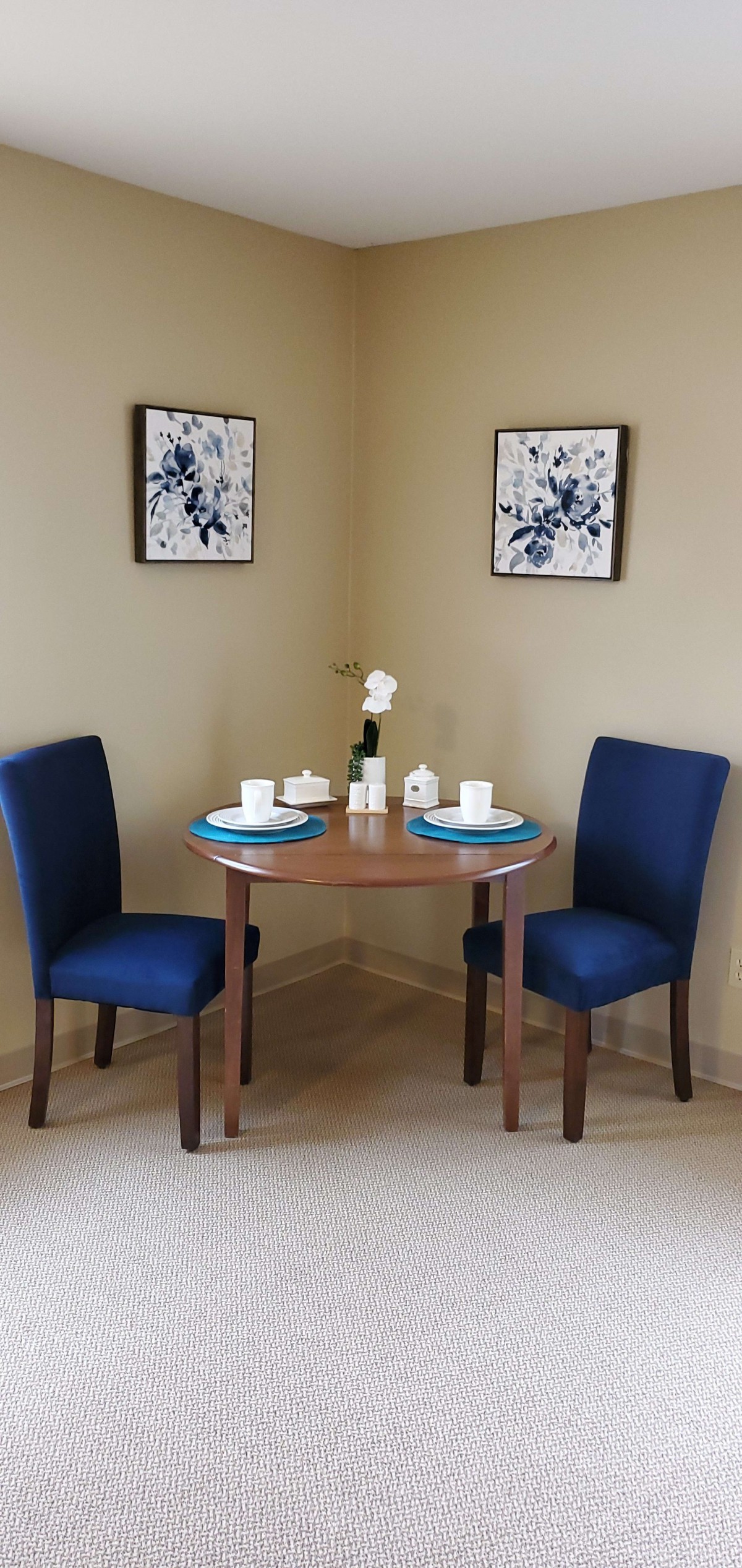 A cozy dining nook with a small round table set for two, featuring blue upholstered chairs and floral wall art.