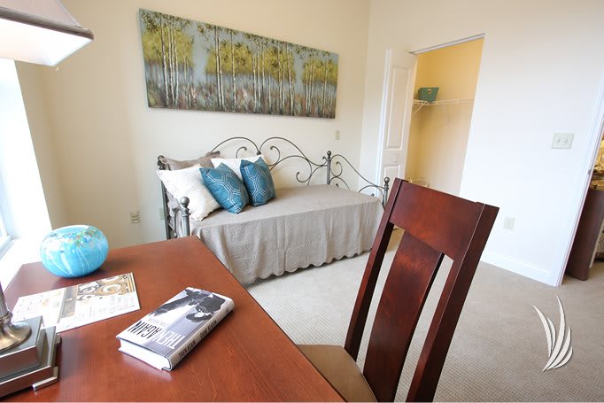 A cozy bedroom features a daybed with decorative pillows and a wooden desk chair, complemented by wall art.