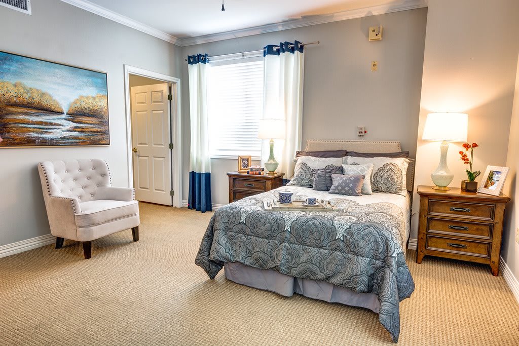 A cozy and well-decorated bedroom featuring a comfortable bed with patterned bedding, a nightstand with a lamp, an armchair, and a window with curtains.