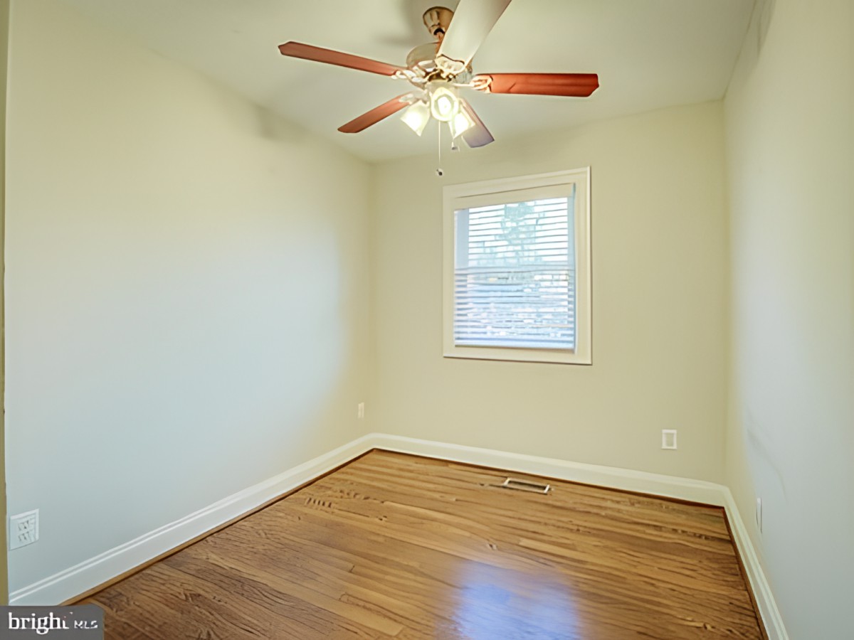 An empty room with hardwood flooring, featuring a ceiling fan and a window providing natural light.