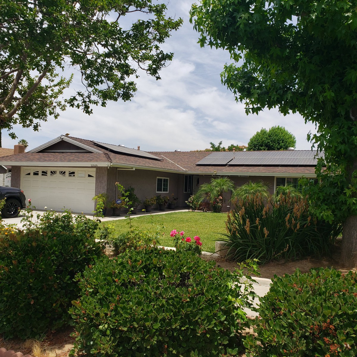 A suburban house with solar panels on the roof, surrounded by a landscaped front yard and trees.