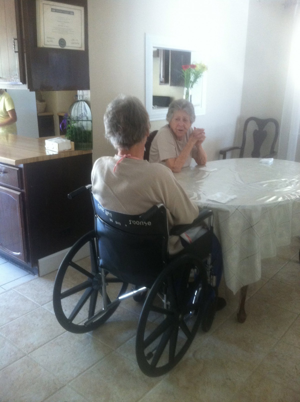 Two elderly women sitting at a table, one in a wheelchair, engaged in conversation.