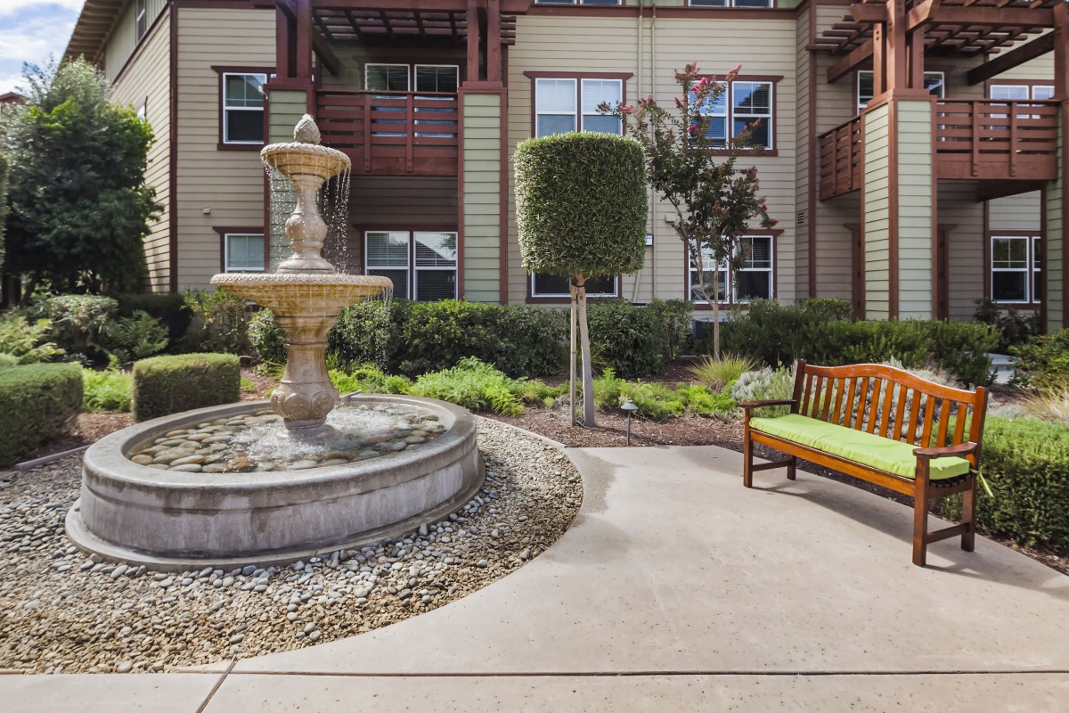 A serene courtyard with a fountain and bench surrounded by lush landscaping in an apartment complex.