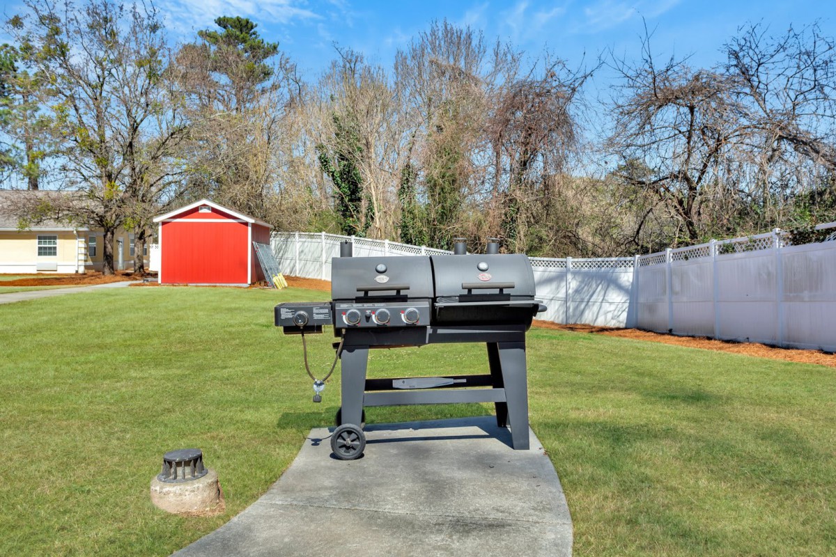 A dual-chamber grill stands in a sunny backyard, surrounded by grass and a white fence.