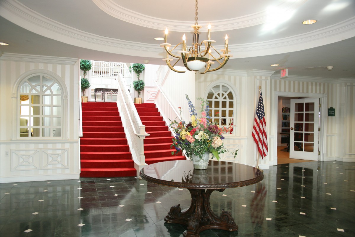 An elegant lobby featuring a red-carpeted staircase, a chandelier, and a central floral arrangement beside the American flag.