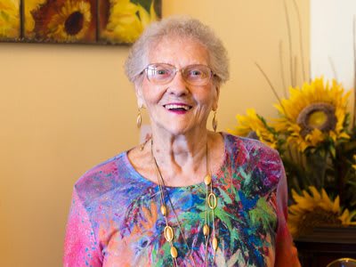 An elderly woman wearing a colorful blouse and glasses, smiling indoors with sunflowers in the background.