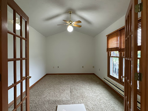 A spacious, empty room with a ceiling fan and large windows, featuring neutral-colored walls and carpet.