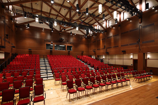 An empty auditorium with red chairs arranged in rows, featuring a wooden interior and stage lighting.