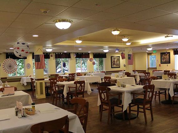 An empty restaurant dining room with tables and chairs neatly arranged, featuring flags and elegant lighting.