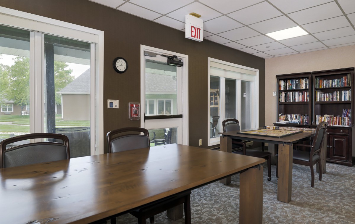 A well-lit meeting room with conference tables and chairs, featuring large windows and an exit sign.
