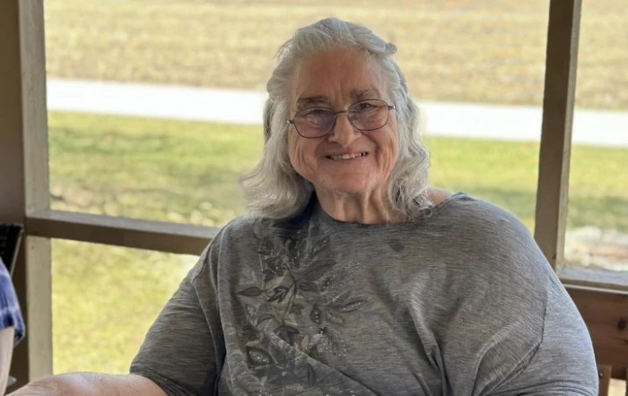 An elderly woman with gray hair and glasses is smiling while seated outdoors.