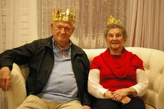 An elderly couple wearing crowns, sitting happily indoors.