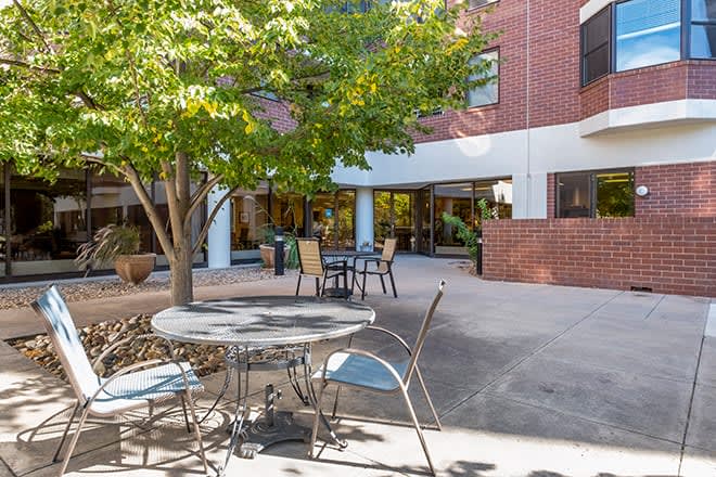 A cozy outdoor patio area with tables and chairs, surrounded by a brick building and shaded by trees.