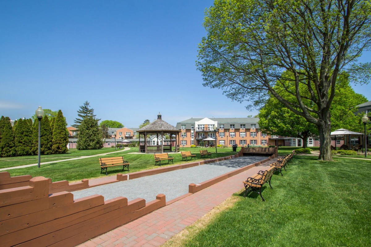 A scenic park area with benches, a gazebo, and a pathway surrounded by greenery and buildings in the background.