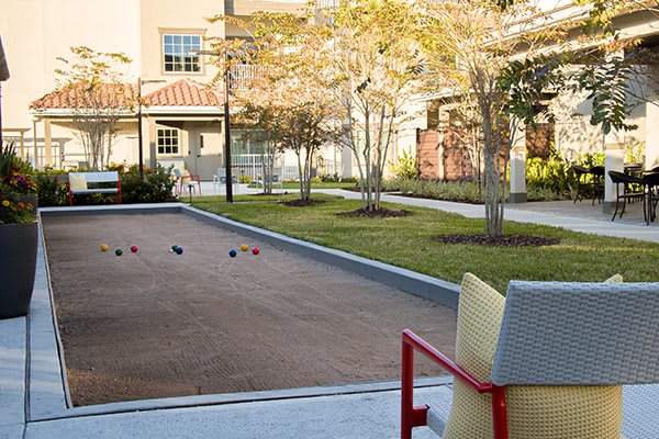 An outdoor bocce court with nearby seating, surrounded by a garden and trees in front of a building.