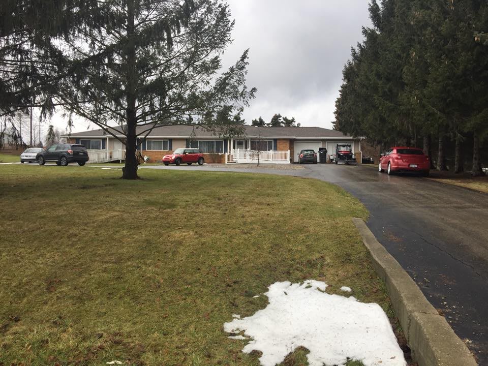 A suburban house with a large lawn and driveway, surrounded by trees on an overcast day, with some snow patches visible.