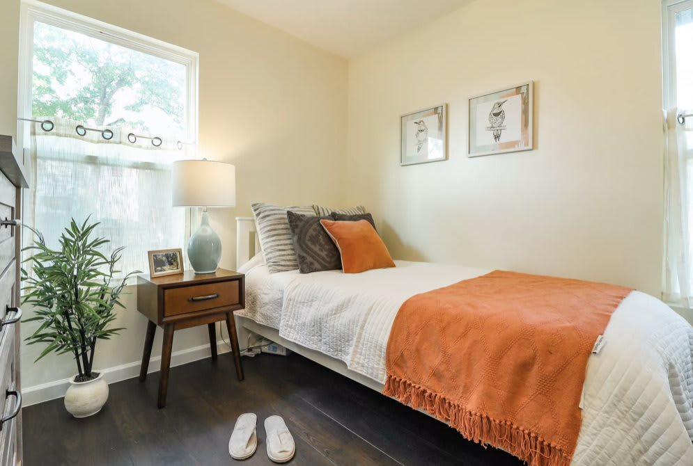 A cozy, minimalist bedroom featuring a neatly made bed with orange accents, a bedside table with a lamp, and a window letting in natural light.