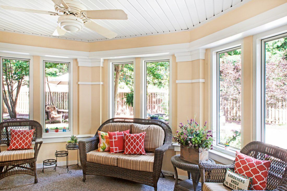 A cozy sunroom with large windows, featuring a rattan sofa, colorful cushions, and decorative plants.