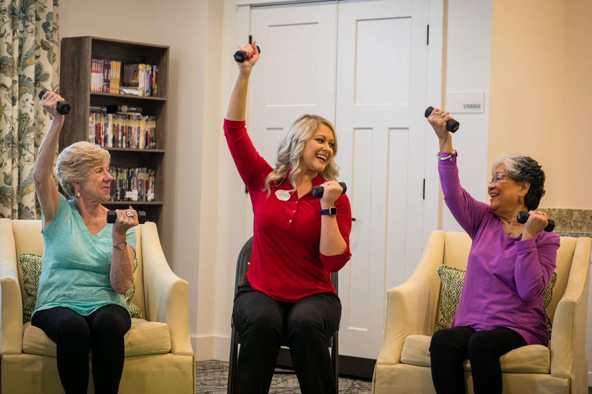 A group of seniors participates in a cheerful exercise session using dumbbells indoors.