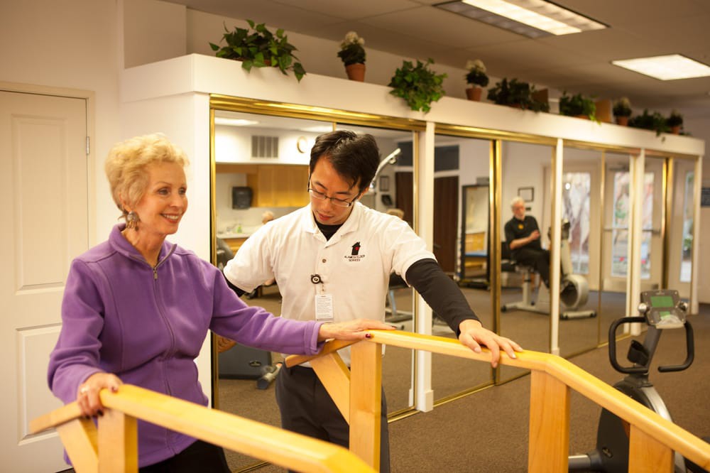 A therapist assists an elderly woman with rehabilitation exercises using parallel bars.