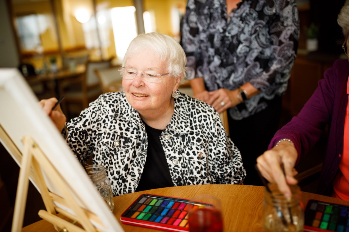 An elderly woman smiles as she participates in a painting activity with others.
