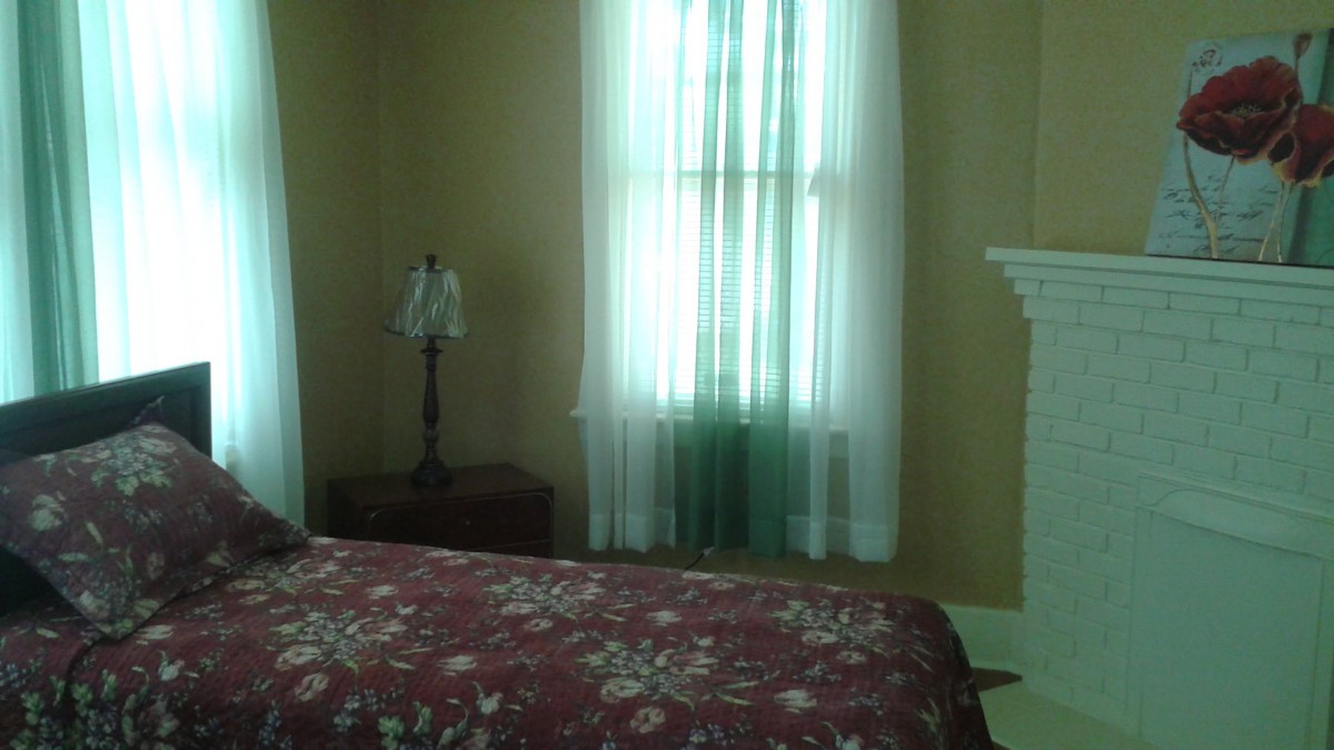 A cozy bedroom featuring a floral bedspread, a window with sheer curtains, and a lamp on the nightstand.