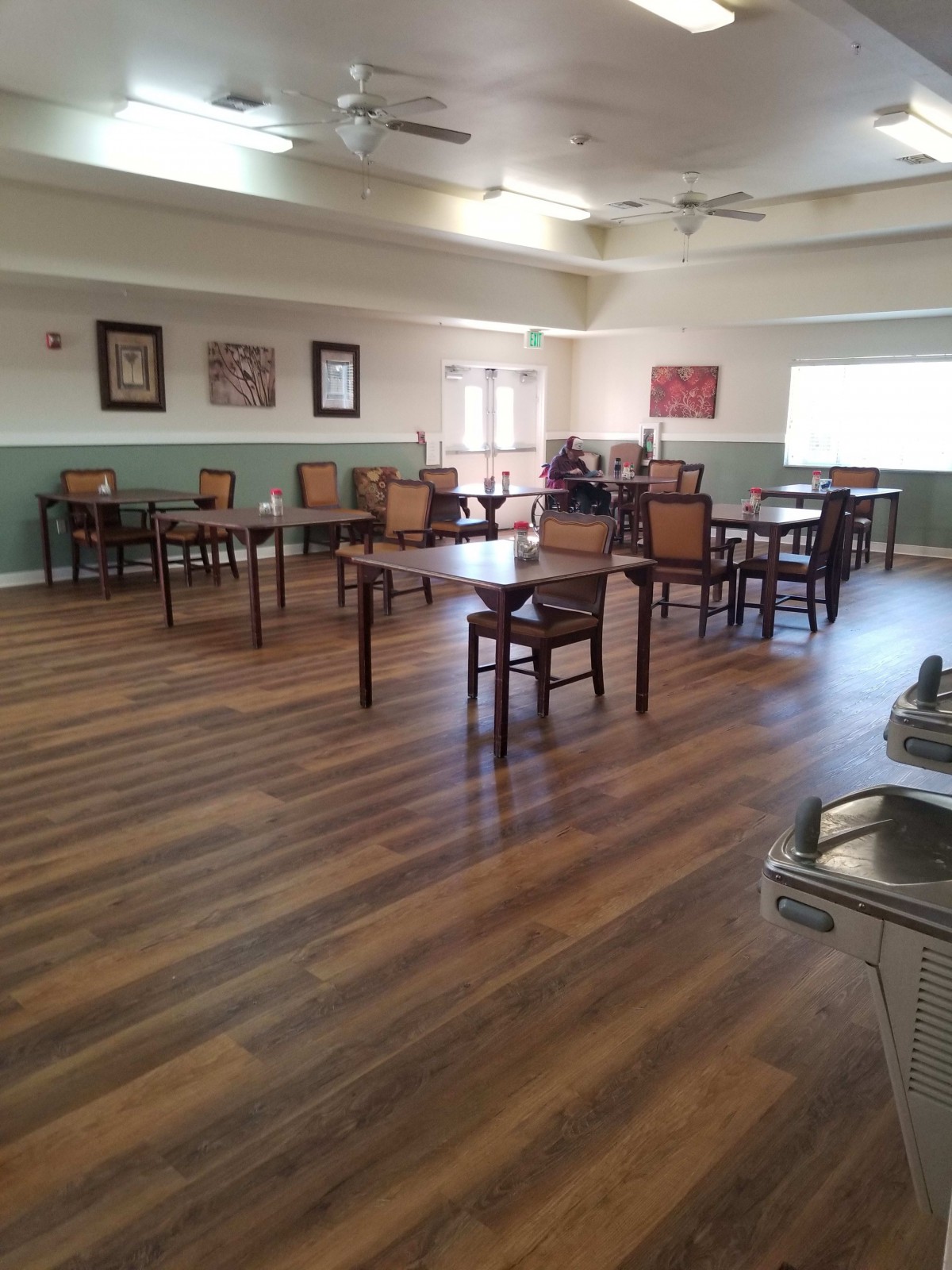 An empty dining room with wooden floors, featuring tables and chairs, and a ceiling fan overhead.