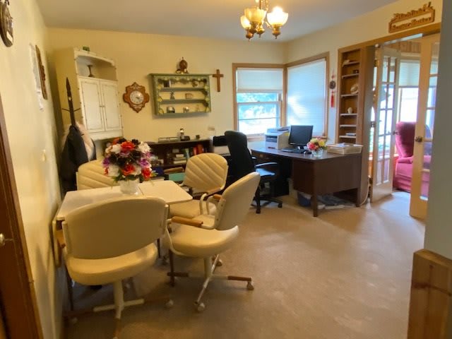 A cozy home office with a desk, bookshelf, and chairs arranged around a table, accented by natural light from the window.