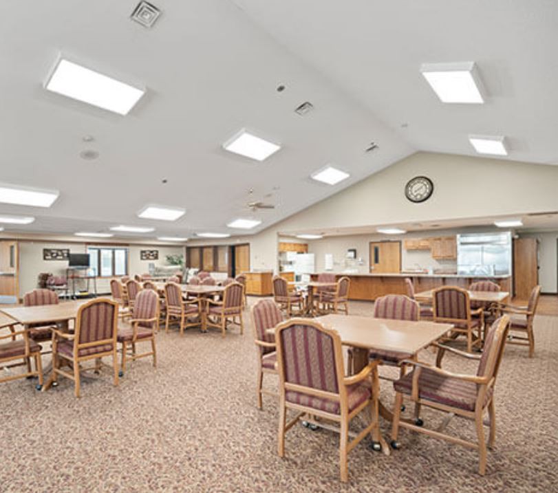 A spacious dining area with multiple tables and chairs, featuring bright overhead lighting and a carpeted floor.