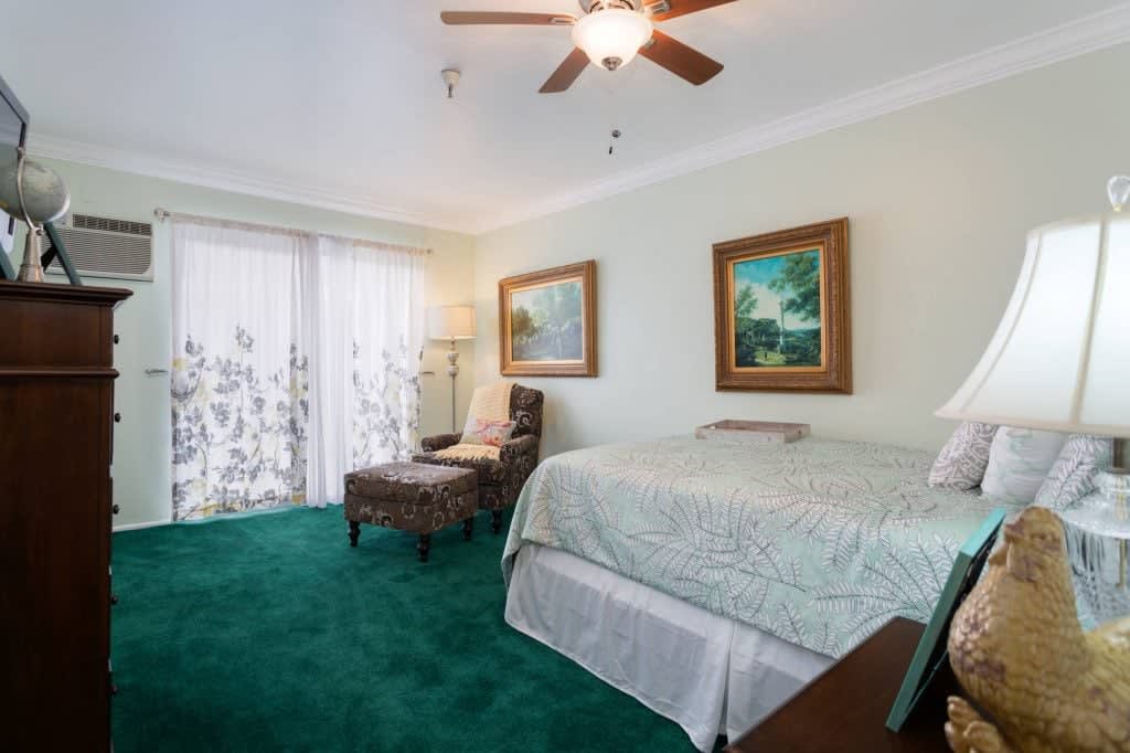 A cozy bedroom with a green carpet, featuring a bed, paintings on the wall, and a ceiling fan.