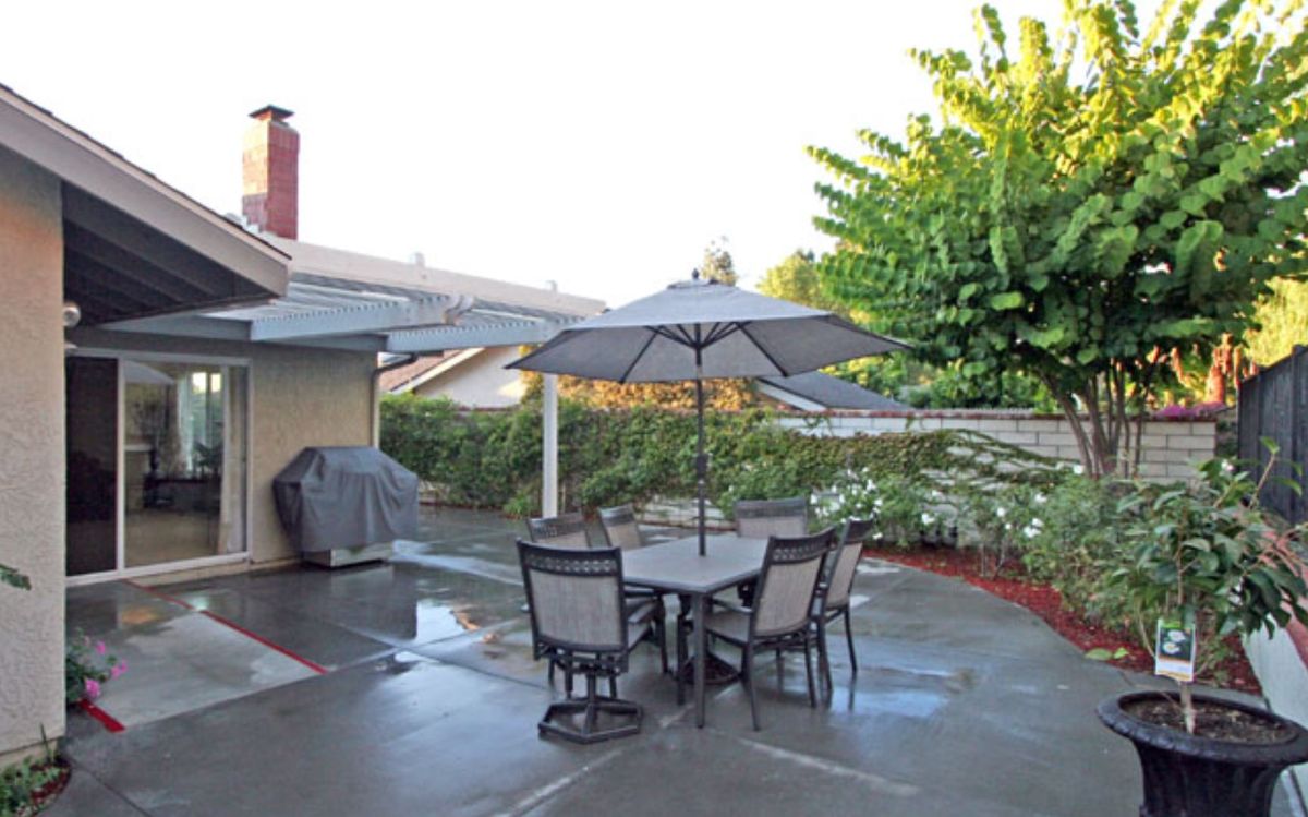 A well-maintained backyard patio with a dining set under an umbrella, surrounded by greenery and featuring a barbecue grill.
