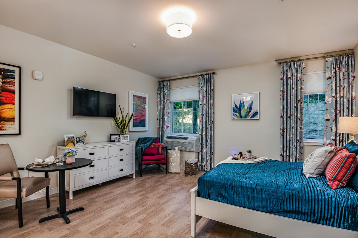 A modern bedroom with blue bedding, a dresser, and a television, complemented by decorative curtains and wall art.