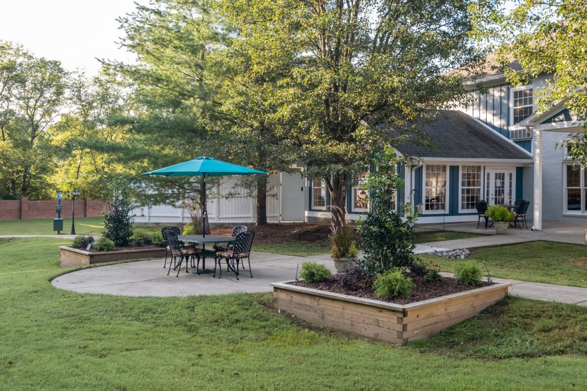 A peaceful backyard garden with a patio set under a blue umbrella, surrounded by lush greenery and a small house in the background.