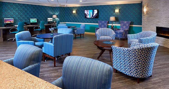 A modern lounge area featuring comfortable seating with blue and patterned chairs, wooden tables, a television on the wall, and ambient lighting from lamps.