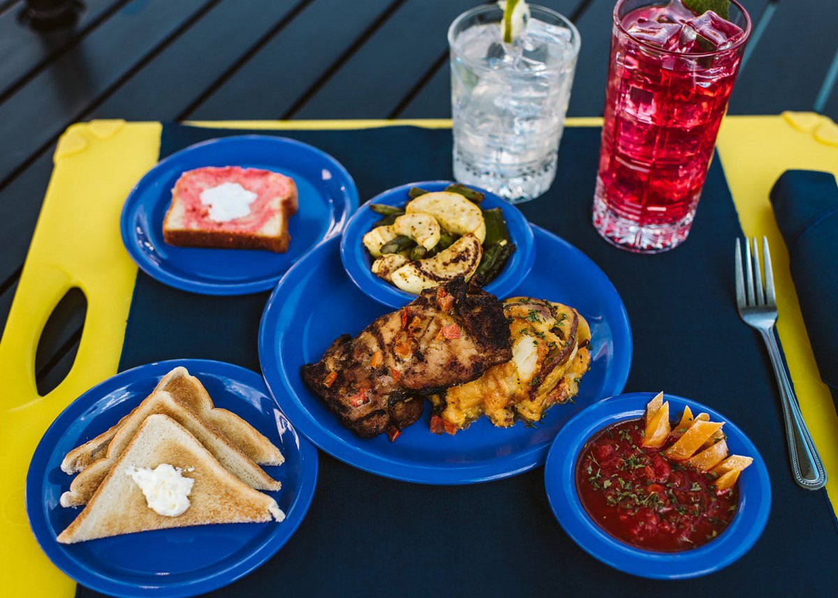 A colorful meal featuring grilled chicken, toast, soup, pickles, and drinks served on blue plates.