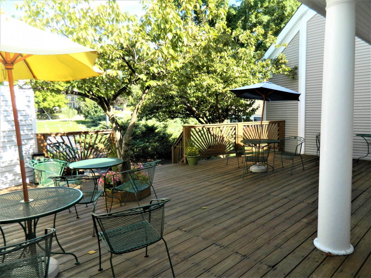 A sunny backyard deck with outdoor seating and umbrellas surrounded by greenery.