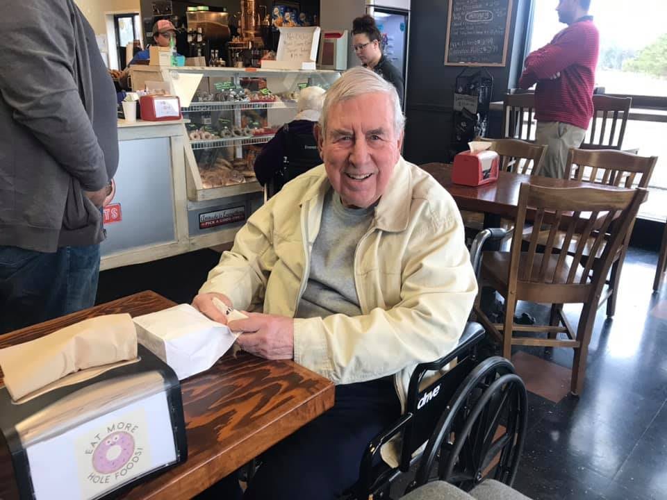 An elderly man in a wheelchair is smiling while seated at a cafe table.