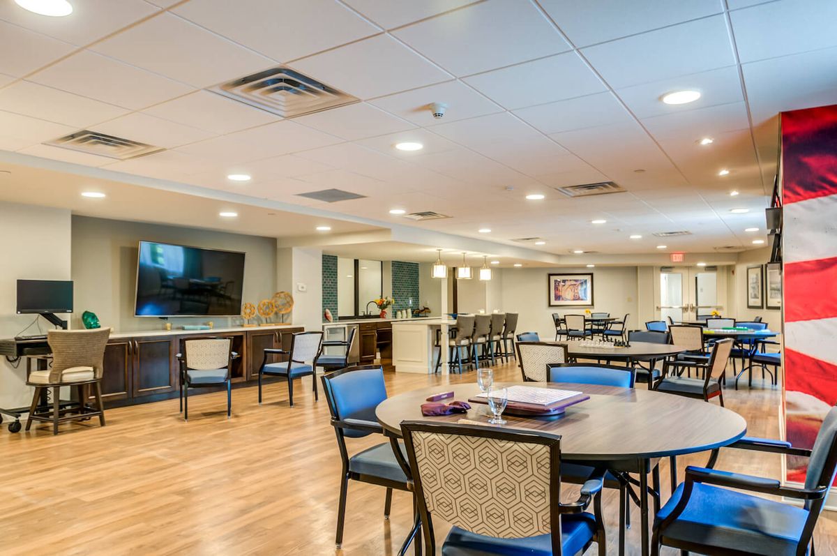 A spacious and well-lit dining area with multiple tables and chairs, featuring a large TV screen on the wall.