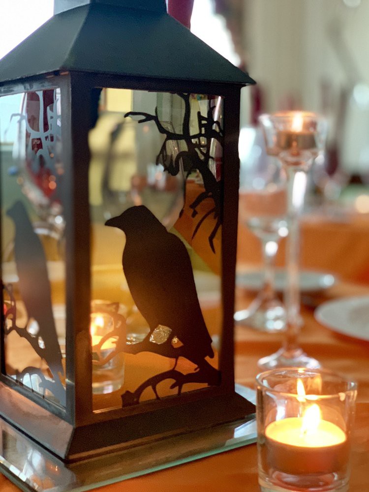 A decorative lantern with a crow silhouette illuminated by candles, set on a dining table.
