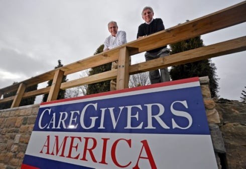 Two people standing behind a 'CareGivers America' sign outdoors.