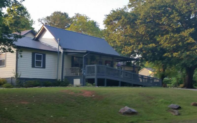 A cozy house with a porch, surrounded by trees and a grassy lawn under the sunlight.