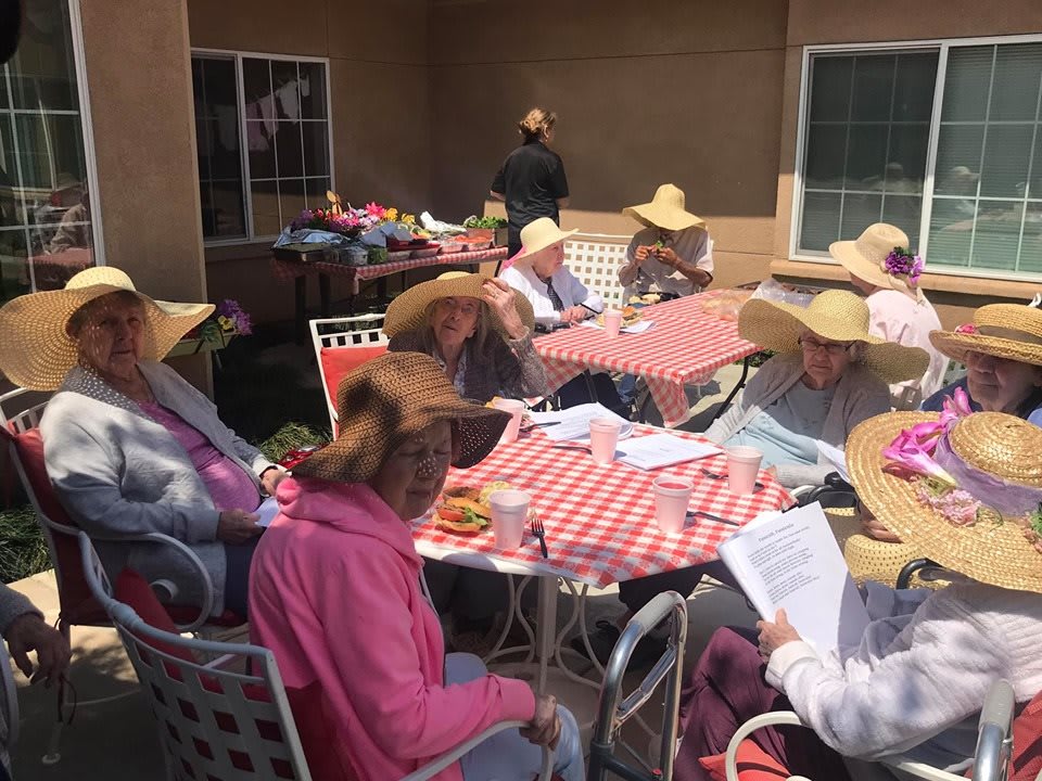 A group of people wearing wide-brimmed hats sit around tables with checkered tablecloths in an outdoor setting.