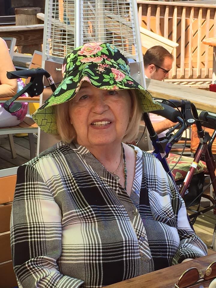 An elderly woman wearing a floral hat and plaid shirt sits outside on a sunny day.